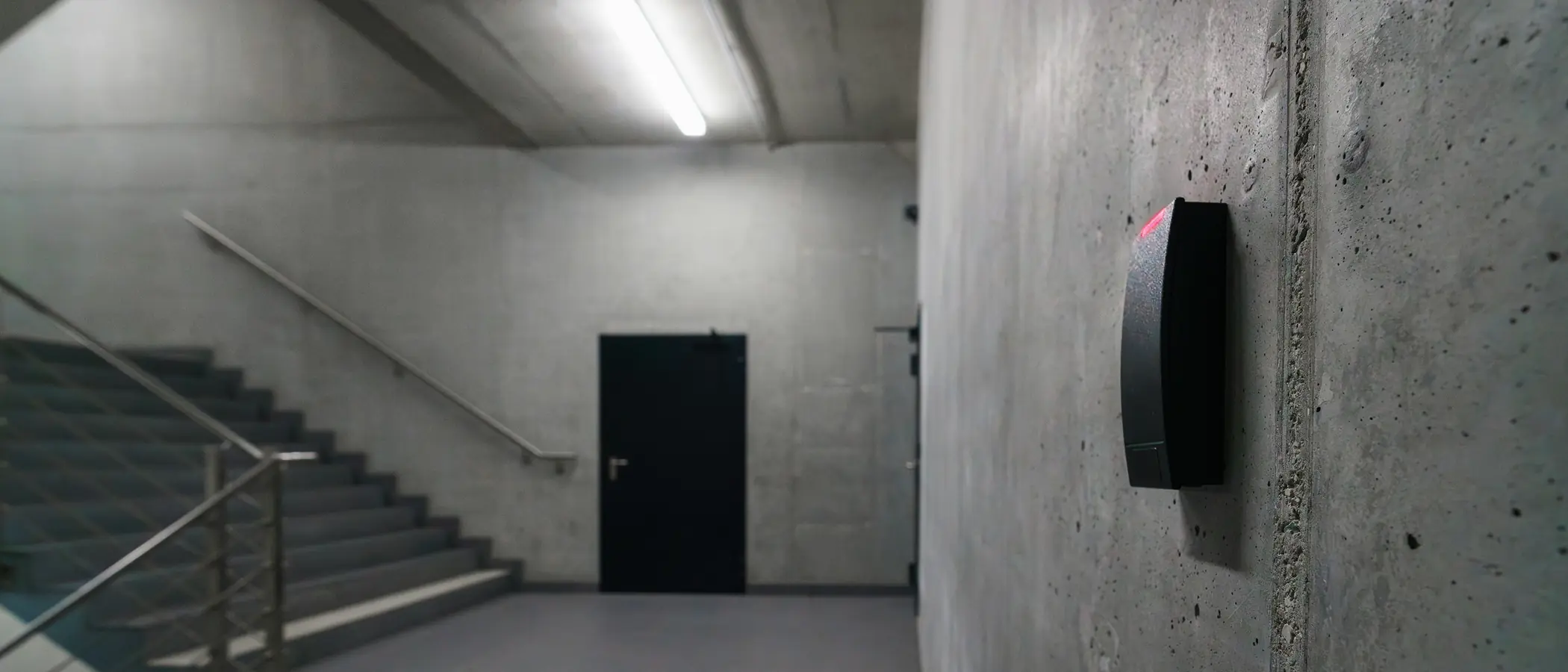 A door-access reader showing a red light on a concrete wall, with a closed door at the end of the corridor.